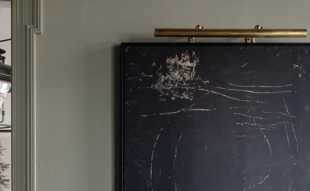 Brass picture light above a large black abstract painting on a plain sage wall